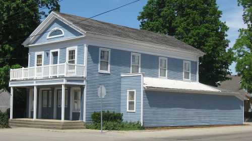 A two-story blue house with a porch, located at a street corner, surrounded by trees and greenery.