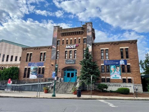 Historic brick building with banners for the Museum of Science and Technology, surrounded by trees and a cloudy sky.