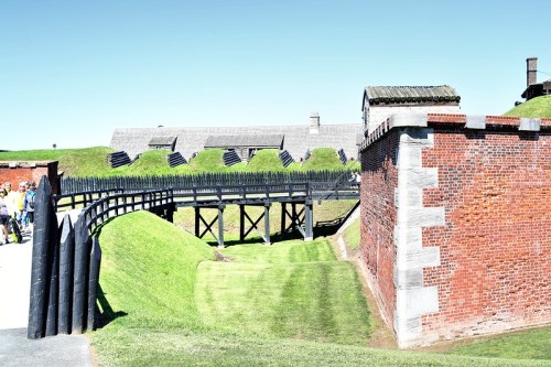 A historic fort with a brick wall, wooden bridge, and grassy mounds under a clear blue sky.