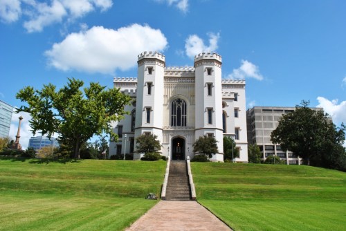 A historic white castle-like building with towers, set on a green lawn under a blue sky with fluffy clouds.