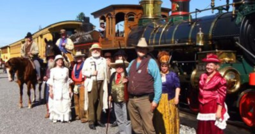 A group of people in historical costumes poses in front of a vintage steam train and colorful carriages.