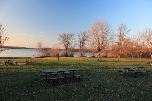 A serene lakeside park at sunset, featuring picnic tables and bare trees along the water's edge.