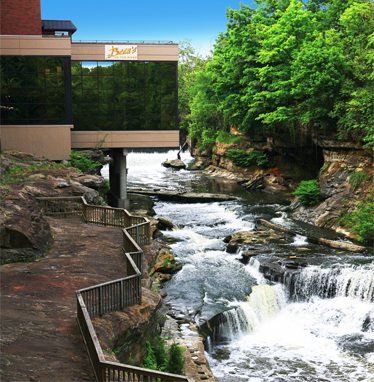 A scenic view of a river with waterfalls, alongside a modern building and lush green trees.