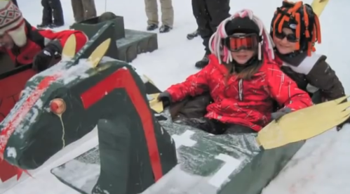 Children in colorful winter gear ride a homemade sled shaped like a horse, surrounded by snow and other kids.