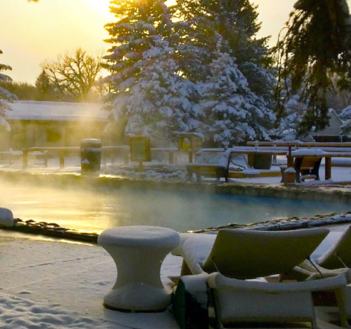 A serene winter scene with a steaming pool surrounded by snow-covered trees and a golden sunrise.