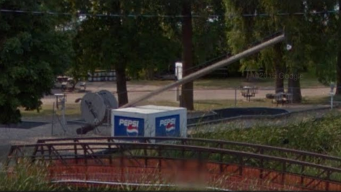 A Pepsi-branded cart is positioned near a roller coaster in a park setting, surrounded by trees and picnic tables.