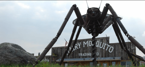 A large metal mosquito sculpture stands in front of a rustic trading post with a sign reading "Mary Mosquito Trading Co."
