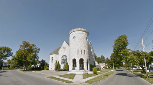 A large stone church with a round tower, surrounded by trees and a clear blue sky, located at a street corner.