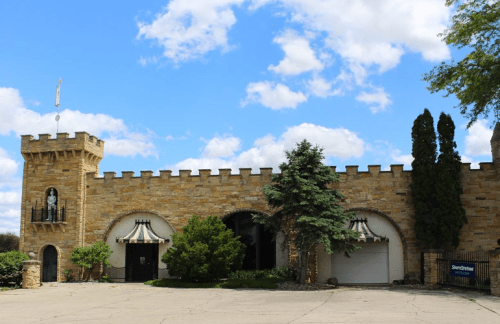 A stone castle-like building with a turret, surrounded by trees and a blue sky with clouds.