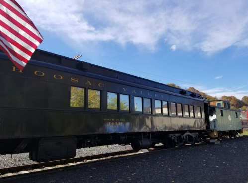A vintage train car labeled "Hoosac Valley" with an American flag in the foreground against a clear blue sky.
