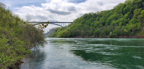 A serene river scene with lush green hills and a bridge spanning across the water under a cloudy sky.