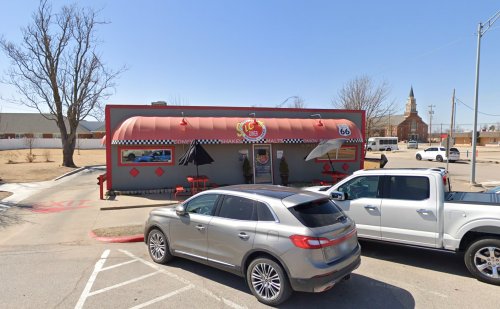 A retro diner with a red awning and checkered accents, parked cars in front, and a church steeple in the background.