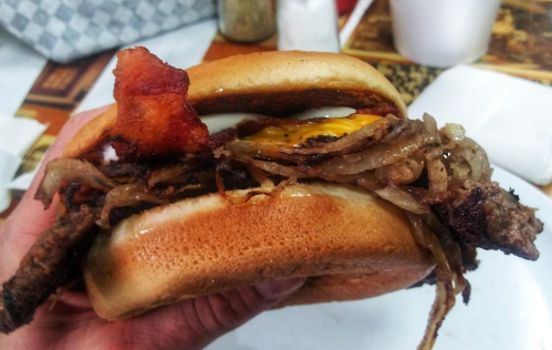 A hand holding a loaded burger with bacon, grilled onions, and melted cheese on a toasted bun.