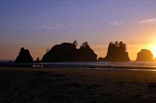 Silhouetted rock formations against a sunset sky, with gentle waves lapping at the sandy beach.