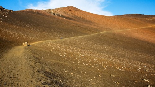 A winding dirt path leads through a barren, rocky landscape with reddish hills under a clear blue sky.