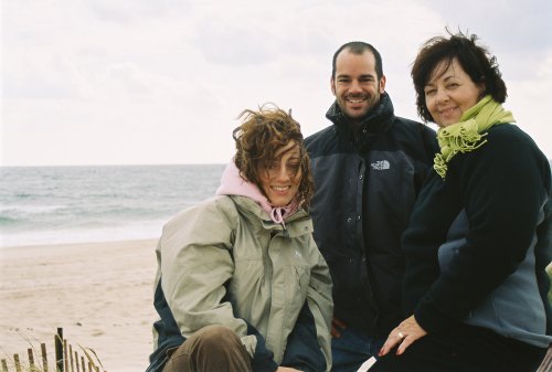Three people smiling on a beach, with waves in the background and cloudy skies above.
