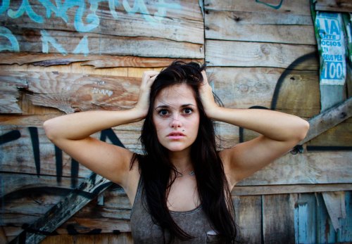 A young woman with long dark hair holds her head in distress against a graffiti-covered wooden wall.