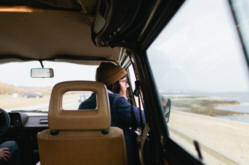 A person wearing a beanie looks out the window of a vehicle, with a scenic view of the beach in the background.