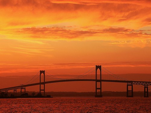 Silhouette of a bridge against a vibrant orange and pink sunset sky over water.