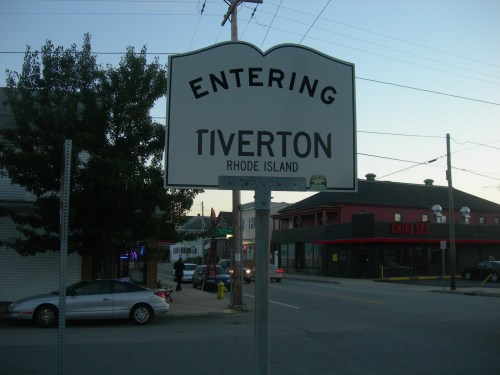 Sign reading "Entering Tiverton, Rhode Island" with a street scene in the background, including cars and buildings.
