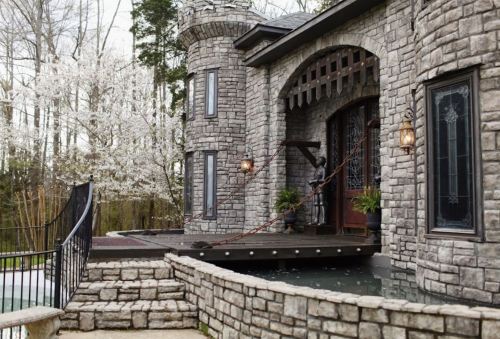 A stone castle-style home with a turret, wooden deck, and blooming trees in the background.