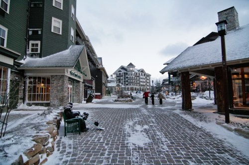 A snowy street in a resort area with shops, people walking, and a person sitting on a bench.