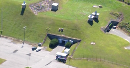Aerial view of a facility with green grass, several structures, and vehicles parked near an entrance.