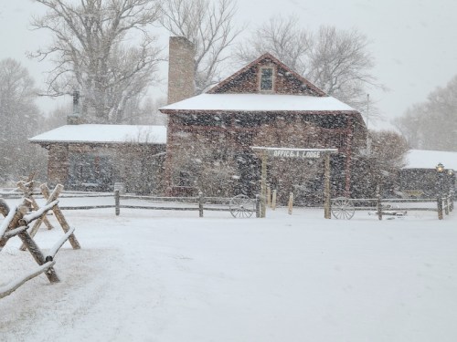 A rustic lodge covered in snow, surrounded by a snowy landscape and trees, with a wooden fence in the foreground.