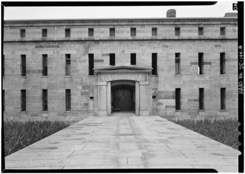 Historic stone building with a central entrance, flanked by narrow windows, leading to a pathway surrounded by grass.