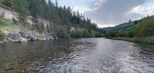 A serene river flows through a lush landscape, surrounded by trees and rocky banks under a cloudy sky.