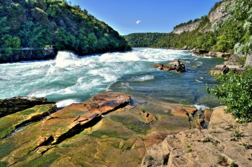 A scenic river flows through rocky terrain, surrounded by lush greenery and steep cliffs under a clear blue sky.