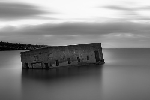A tilted concrete structure partially submerged in calm water, set against a cloudy sky in black and white.
