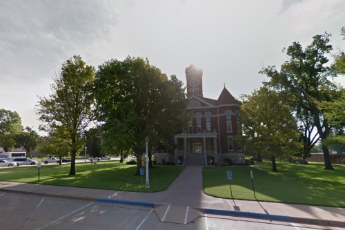 Historic brick courthouse surrounded by trees and green lawn, with a clock tower visible against a cloudy sky.