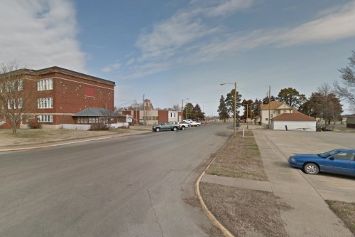 A quiet street scene featuring a brick building, parked cars, and a clear sky in a small town.