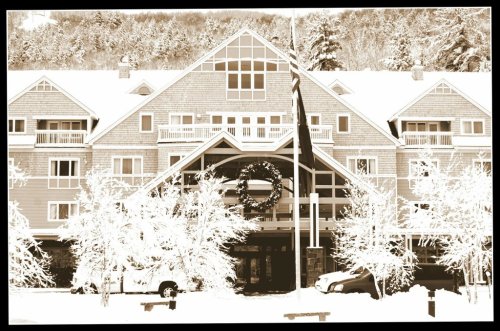 A snow-covered hotel entrance with a large wreath, surrounded by trees and a flagpole in winter scenery.