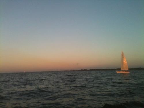 A sailboat glides across calm waters at sunset, with a gradient sky transitioning from orange to blue.