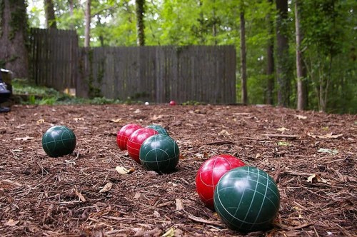 A bocce ball court in a wooded area, featuring red and green balls on a brown mulch surface.