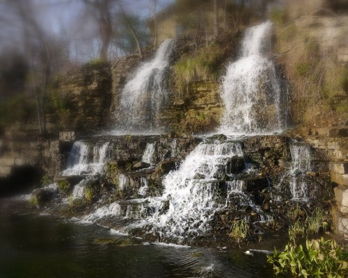 A serene waterfall cascading over rocky ledges into a calm pool, surrounded by greenery and soft sunlight.