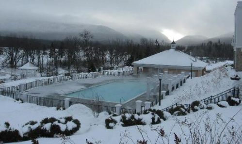 A snowy landscape featuring a pool surrounded by snow-covered ground and mountains in the background under a cloudy sky.