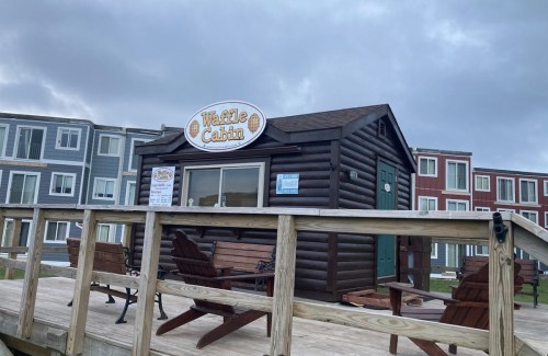 A small wooden cabin with a sign reading "Waffle Cabin," surrounded by picnic tables and a cloudy sky.