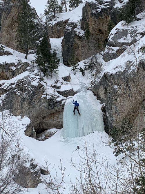 A climber ascends a frozen waterfall surrounded by rocky cliffs and snow-covered trees.