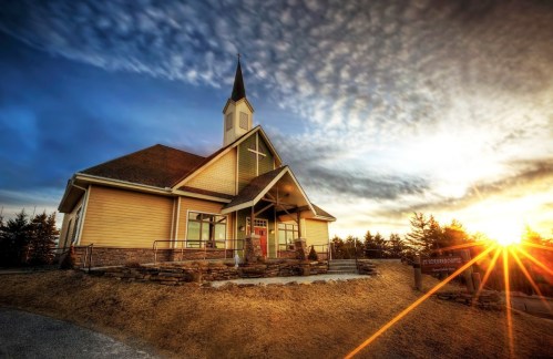 A charming church with a steeple, set against a dramatic sunset and cloudy sky, surrounded by trees.