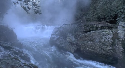 A misty river flows over rocks, surrounded by lush greenery and steam rising from the water.