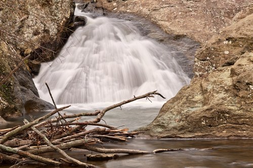 A serene waterfall cascades over rocks, surrounded by branches and a calm river.