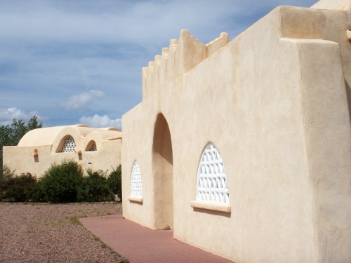 Adobe-style buildings with arched windows and a sandy exterior, set against a blue sky with scattered clouds.