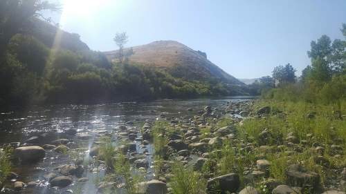 A serene river flows through a rocky landscape, surrounded by lush greenery and hills under a clear blue sky.