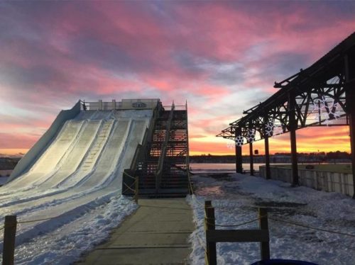 A snowy slide beside a frozen lake at sunset, with vibrant pink and orange skies and a rustic structure nearby.