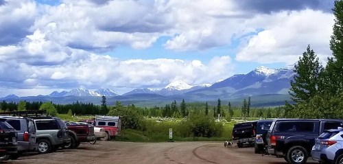 A scenic view of snow-capped mountains under a cloudy sky, framed by parked vehicles in a dirt lot.