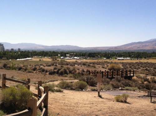 A scenic view of a valley with green trees, distant mountains, and a wooden bridge in the foreground under a clear blue sky.