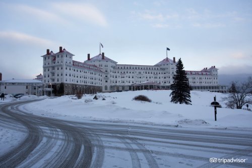 A large white hotel with red roofs surrounded by snow, set against a cloudy sky.